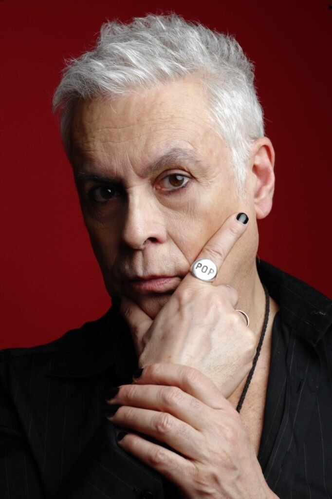 Valentino and the pims en portrait rapproché sur fond rouge. Cheveux blancs courts, eye liner noir façon rock, ongles noir. Chemise noire haute couture et chevalière "pop" sur l'index droit. Rock punk attitude. Generation X.